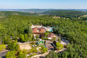 Luftaufnahme eines Campingplatzes im Grünen mit mehreren Gebäuden, Wegen, Stellplätzen und einem kleinen Pool zwischen Bäumen.