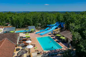 Großer Camping-Pool mit blauen Rutschen, Liegen und Sitzbereich, umgeben von Bäumen und mehreren Gebäuden.