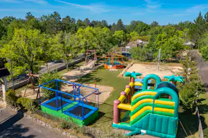 Spielplatz auf dem Campingplatz mit Hüpfburg, Rutsche und blauem Klettergerüst, umgeben von Bäumen und Wegen.