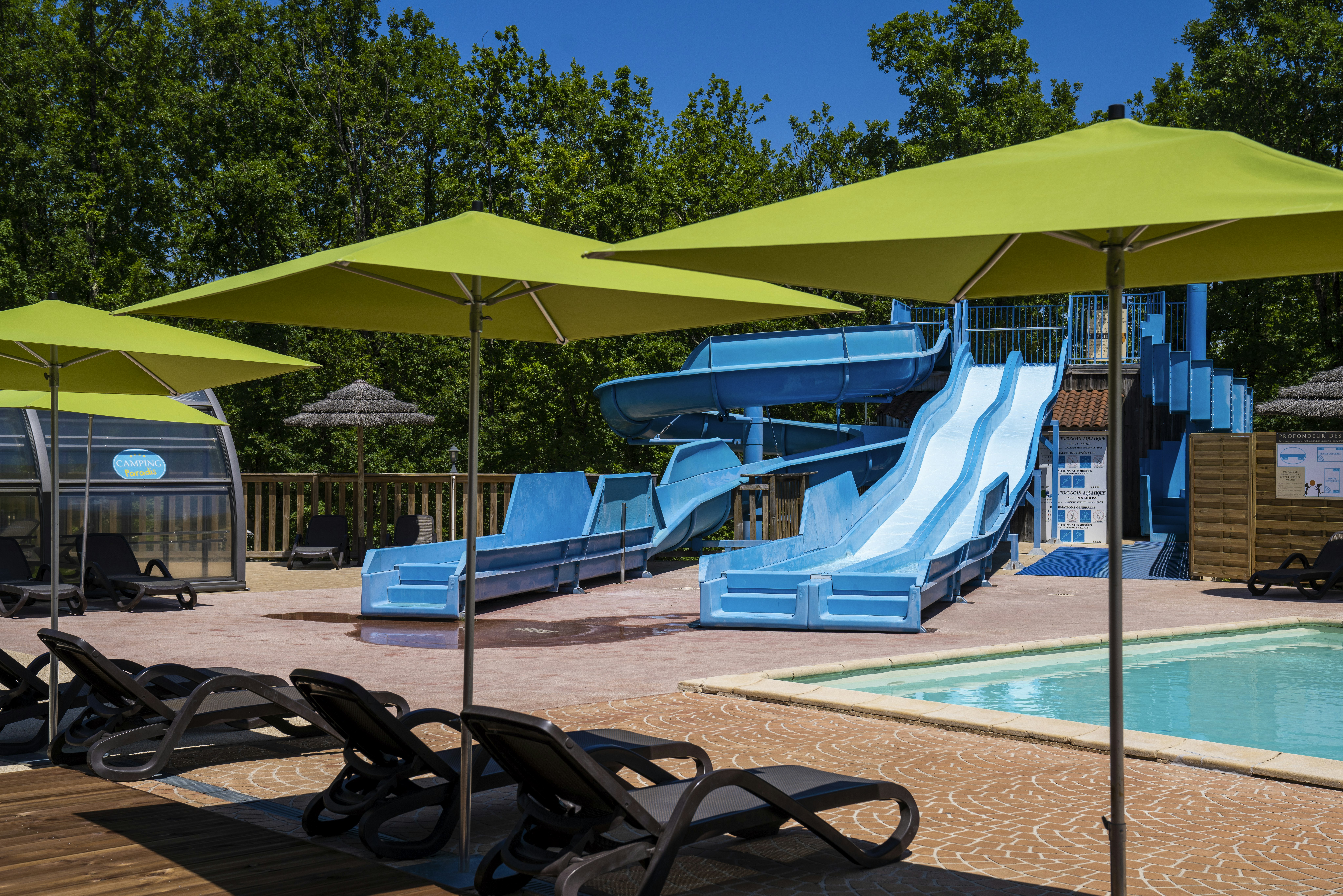 Blauer Pool mit Rutsche, Liegen und Sonnenschirmen auf dem Campingplatz, im Hintergrund Bäume und ein Gebäude.