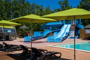 Blauer Pool mit Rutsche, Liegen und Sonnenschirmen auf dem Campingplatz, im Hintergrund Bäume und ein Gebäude.