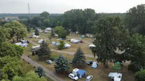 Campingplatz mit Wohnmobilen und Zelten auf Stellplätzen, umgeben von Bäumen und Wegen, Blick von oben auf das Gelände.