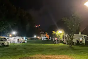 Campingplatz bei Nacht: Wohnwagen und Zelte am beleuchteten Rasen, daneben ein Tisch und Stühle.
