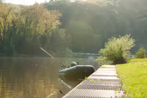 Boot am See neben einem Steg, grünes Ufer mit Bäumen und Gras, ruhiges Wasser im Hintergrund.