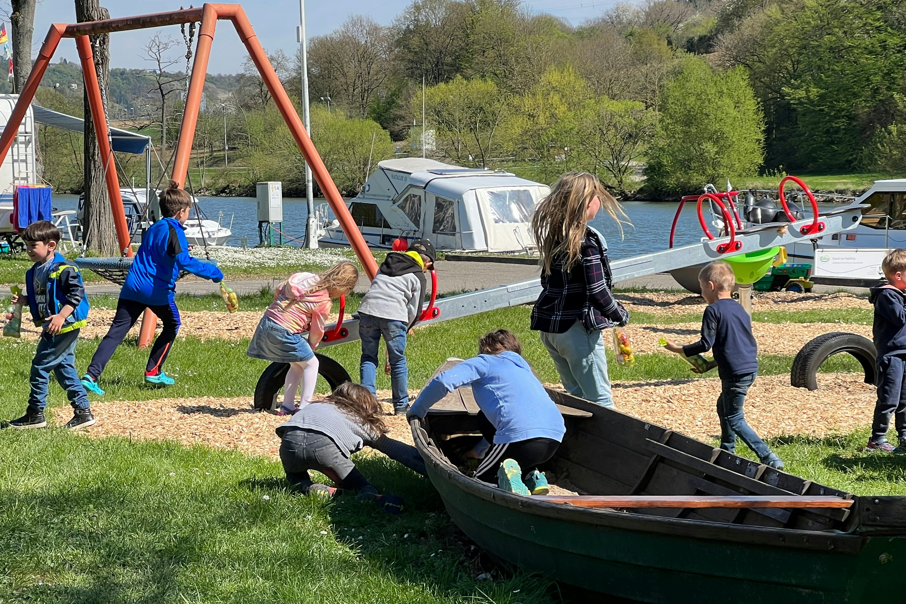 Kinder spielen auf einem Campingplatz-Spielplatz mit einem großen Schubkarren und Schaukelgestell, im Hintergrund Wohnwagen und Zelte.