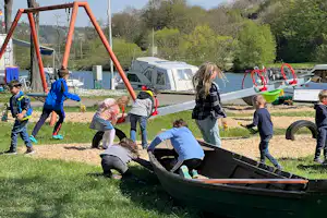 Kinder spielen auf einem Campingplatz-Spielplatz mit einem großen Schubkarren und Schaukelgestell, im Hintergrund Wohnwagen und Zelte.