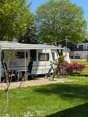 Wohnwagen auf einem Campingplatz mit Vorzelt, Gartenmöbeln und Fahrrad am Stellplatz, umgeben von Bäumen und Rasen.