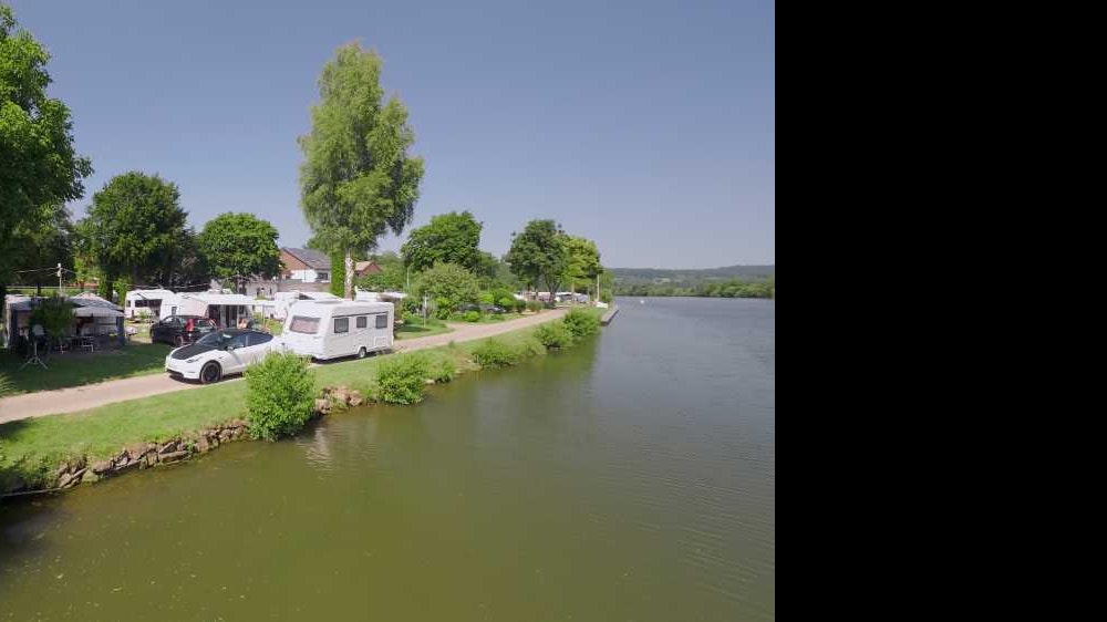 Wohnmobile und Campingplätze am Ufer eines Sees, mit Bäumen, Grasstreifen und einem kleinen Steg am Rand.