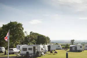 Wohnmobile auf einem Campingplatz, im Vordergrund eine Flagge, dahinter Bäume und Wiesen unter klarem Himmel.