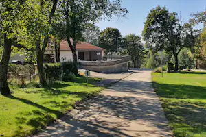 Campingweg zwischen Bäumen und Rasenflächen, mit Gebäuden im Hintergrund und einem Zaun entlang der Strecke.