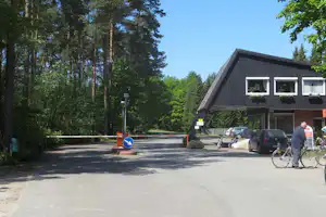 Campinghaus am Parkplatz im Wald, mit Fahrrädern, Autos und Einfahrtsschild vor dem Gebäude.