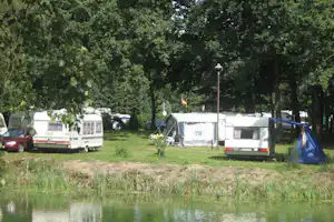 Wohnwagen und Zelte auf einem Campingplatz am See, umgeben von Bäumen und Rasenflächen.