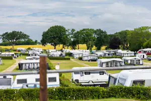 Wohnwagen und Campingbusse auf einem Stellplatz, umgeben von Hecken und Bäumen, mit breitem Weg im Campinggelände.