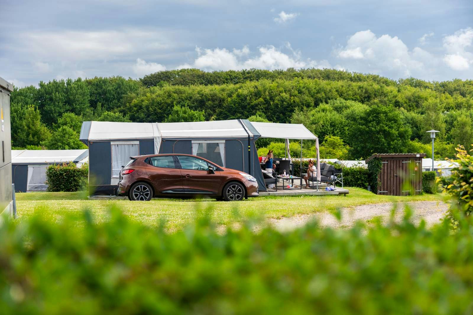 Stellplatz mit Wohnwagen und Auto vor einer modernen Überdachung, Sitzgruppe und Rasenfläche im Campingbereich, umgeben von Bäumen.