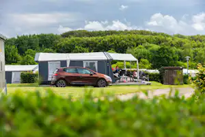Stellplatz mit Wohnwagen und Auto vor einer modernen Überdachung, Sitzgruppe und Rasenfläche im Campingbereich, umgeben von Bäumen.