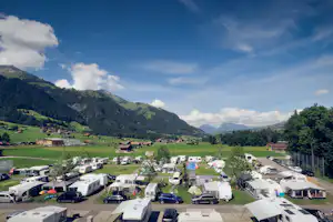 Blick von oben auf einen Campingplatz mit Wohnmobilen und Stellplätzen, umgeben von grünen Bergen und Wäldern.