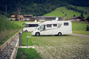 Wohnmobil und Campingbus auf einem Stellplatz mit gepflastertem Weg, Hecken und Wiese, im Hintergrund Gebäude und Berge.