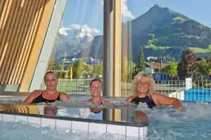 Drei Personen im Pool vor Glasfront, im Hintergrund Berge und Bäume, daneben ein Zaun und Gebäude am Campingplatz.