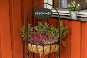 Willkommensschild an einem Metallständer mit Blumen und Kräutern in einem Jute-Sack vor einer roten Holzwand.