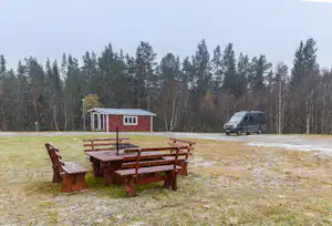 Holz-Tischgruppe auf einem Campingplatz vor einem roten Holzhaus und einem schwarzen Transporter, umgeben von Bäumen.