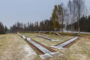 Minigolfanlage mit mehreren Bahnen, roten Begrenzungen und Kiesboden, im Hintergrund Bäume und ein Weg am Campingplatz.
