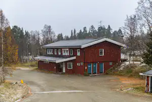 Rotes Campinghaus mit Satteldach, mehreren Fenstern und Eingang, umgeben von Bäumen am Rand eines Campingplatzes.