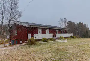 Rotes Ferienhaus mit Holzveranda und weißen Geländern, umgeben von Bäumen und Rasenfläche auf dem Campingplatz.