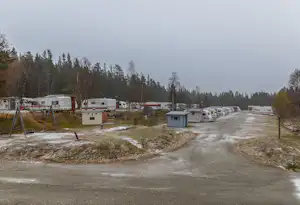Reihe von Wohnwagen und Caravans auf einem Campingplatz, mit Schotterweg, kleinen Gebäuden und Bäumen im Hintergrund.