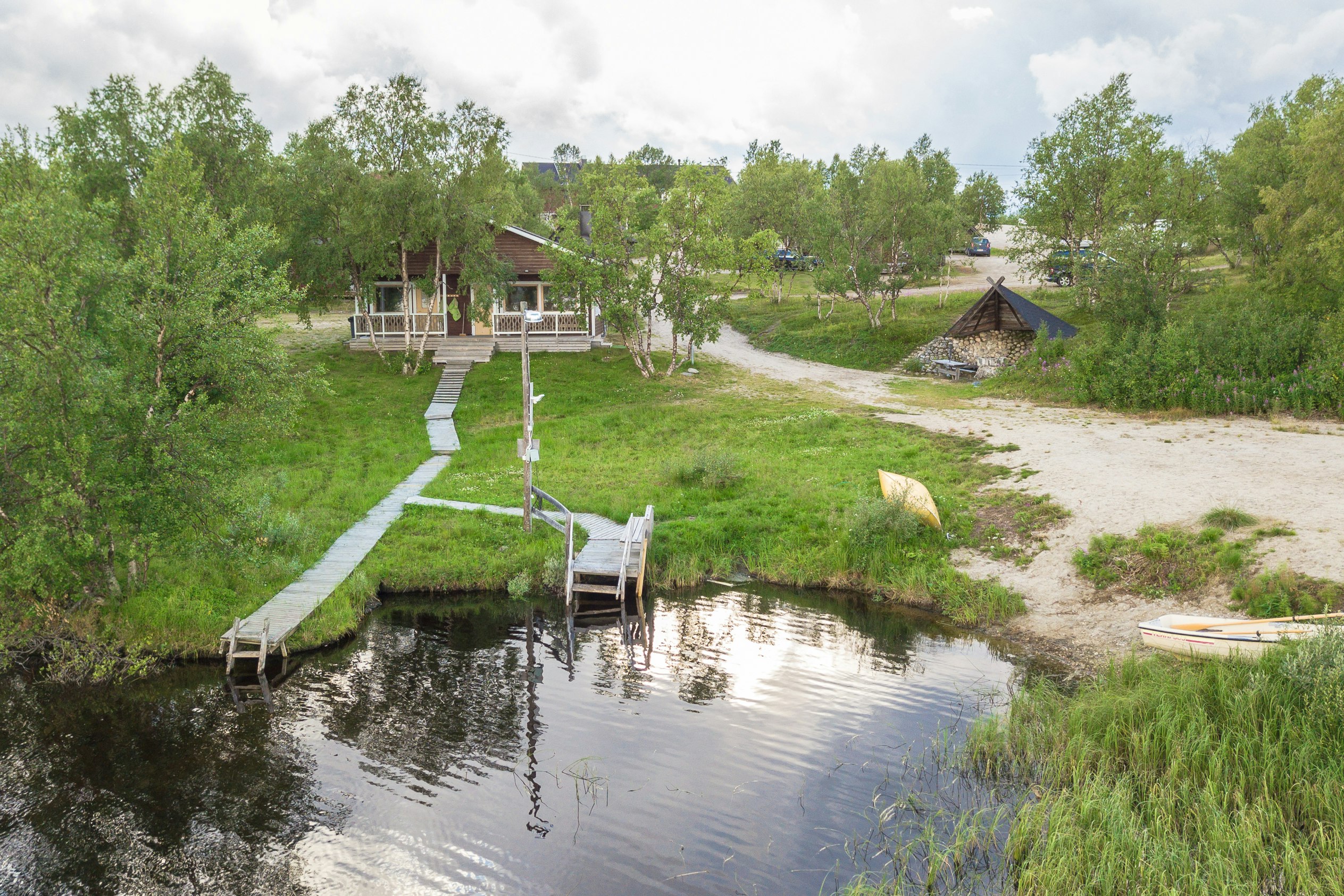 Holzsteg und Sitzbereich am kleinen Wasserbecken im Campingbereich, umgeben von Rasen, Bäumen und Wegen.