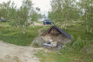 Ein kleines Steinhaus mit Holzschindeldach am Campingplatz, daneben ein Tisch und Sitzbank, umgeben von Bäumen und Wegen.