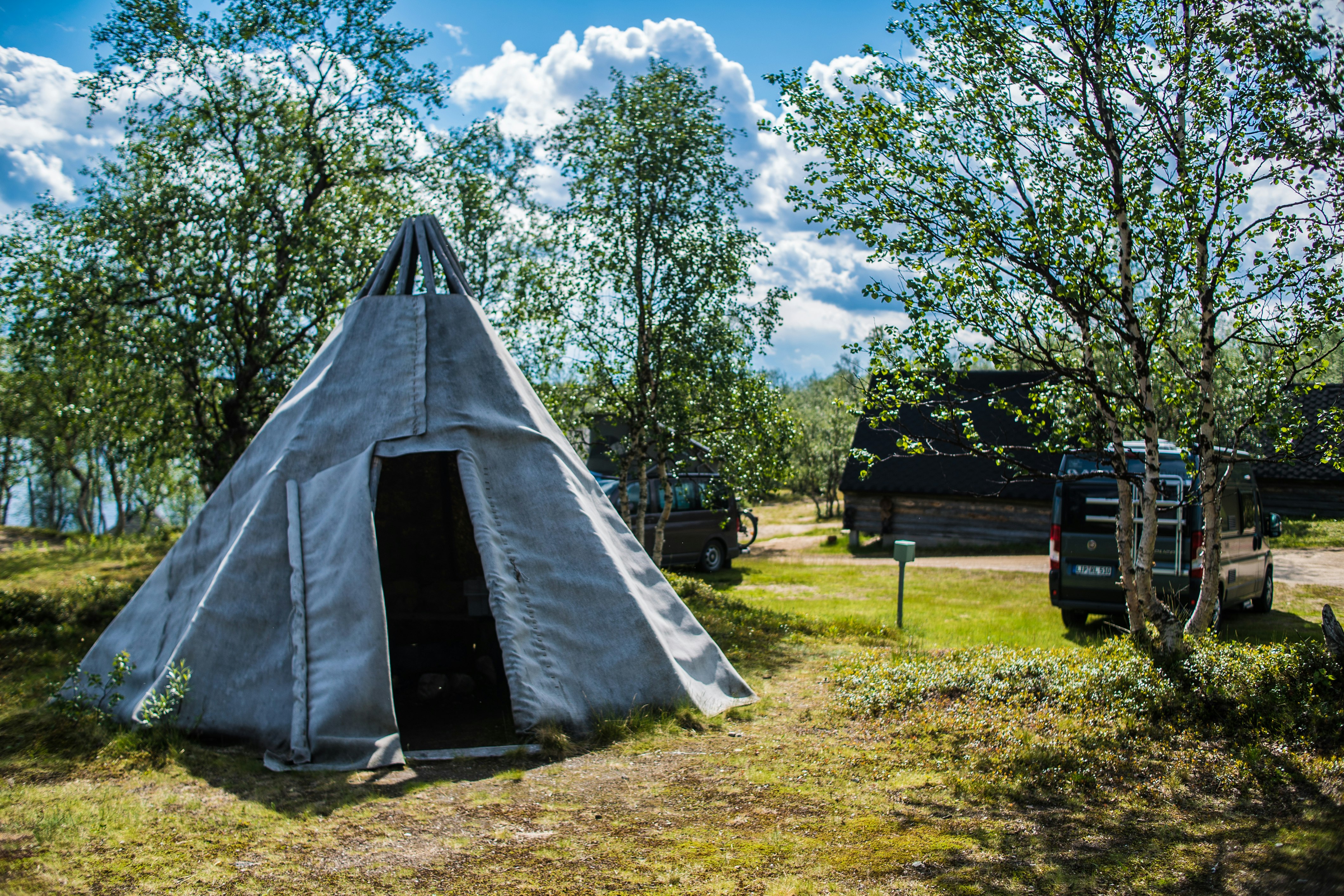 Zelt auf einem grasigen Campingplatz, umgeben von Bäumen; im Hintergrund stehen Fahrzeuge und ein kleiner Weg.