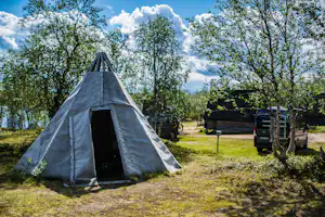 Zelt auf einem grasigen Campingplatz, umgeben von Bäumen; im Hintergrund stehen Fahrzeuge und ein kleiner Weg.