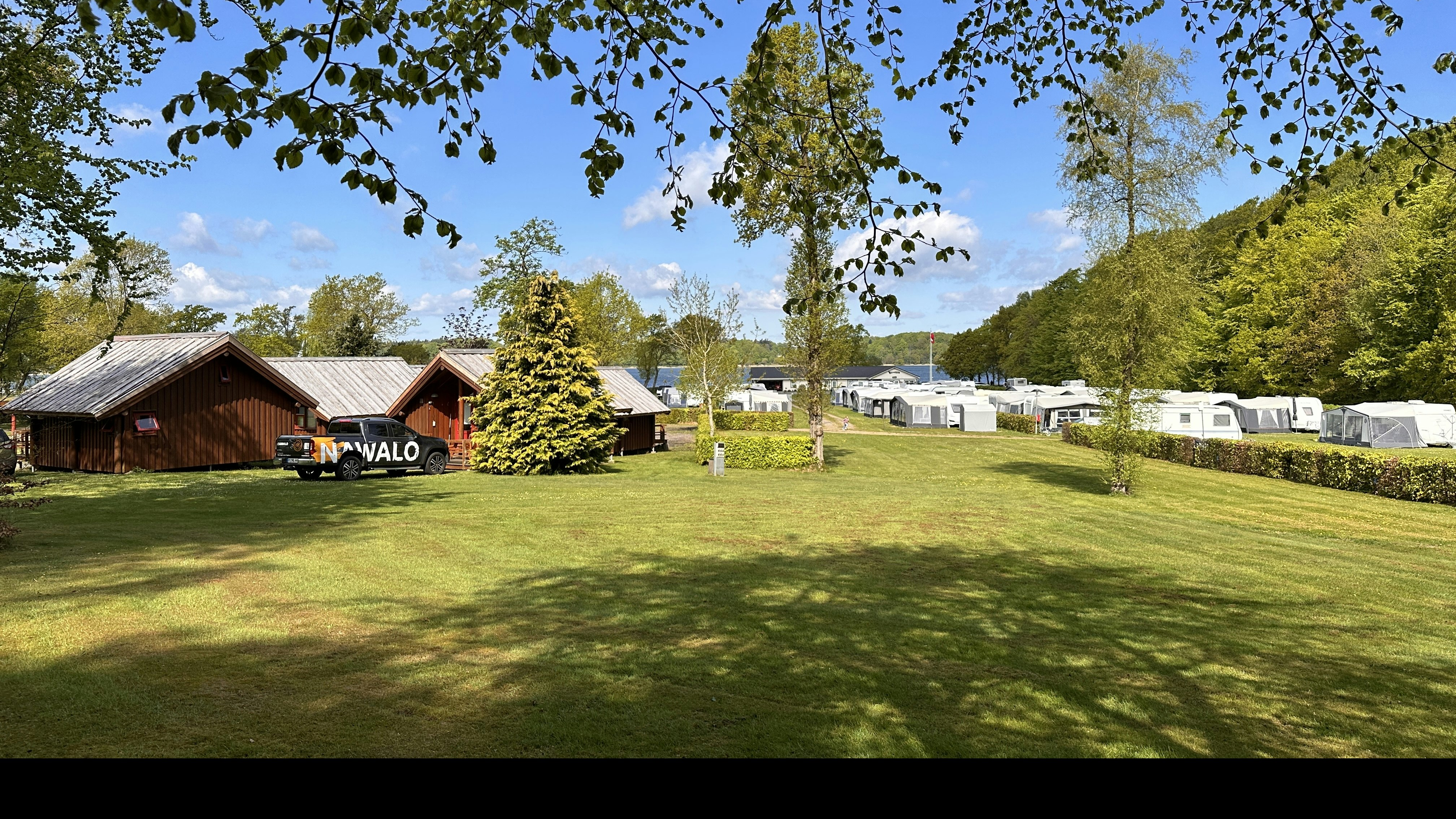 Campingplatz mit Rasenfläche, Holzhäusern und Wohnwagen, im Hintergrund Bäume und ein Seeufer.