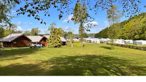 Campingplatz mit Rasenfläche, Holzhäusern und Wohnwagen, im Hintergrund Bäume und ein Seeufer.