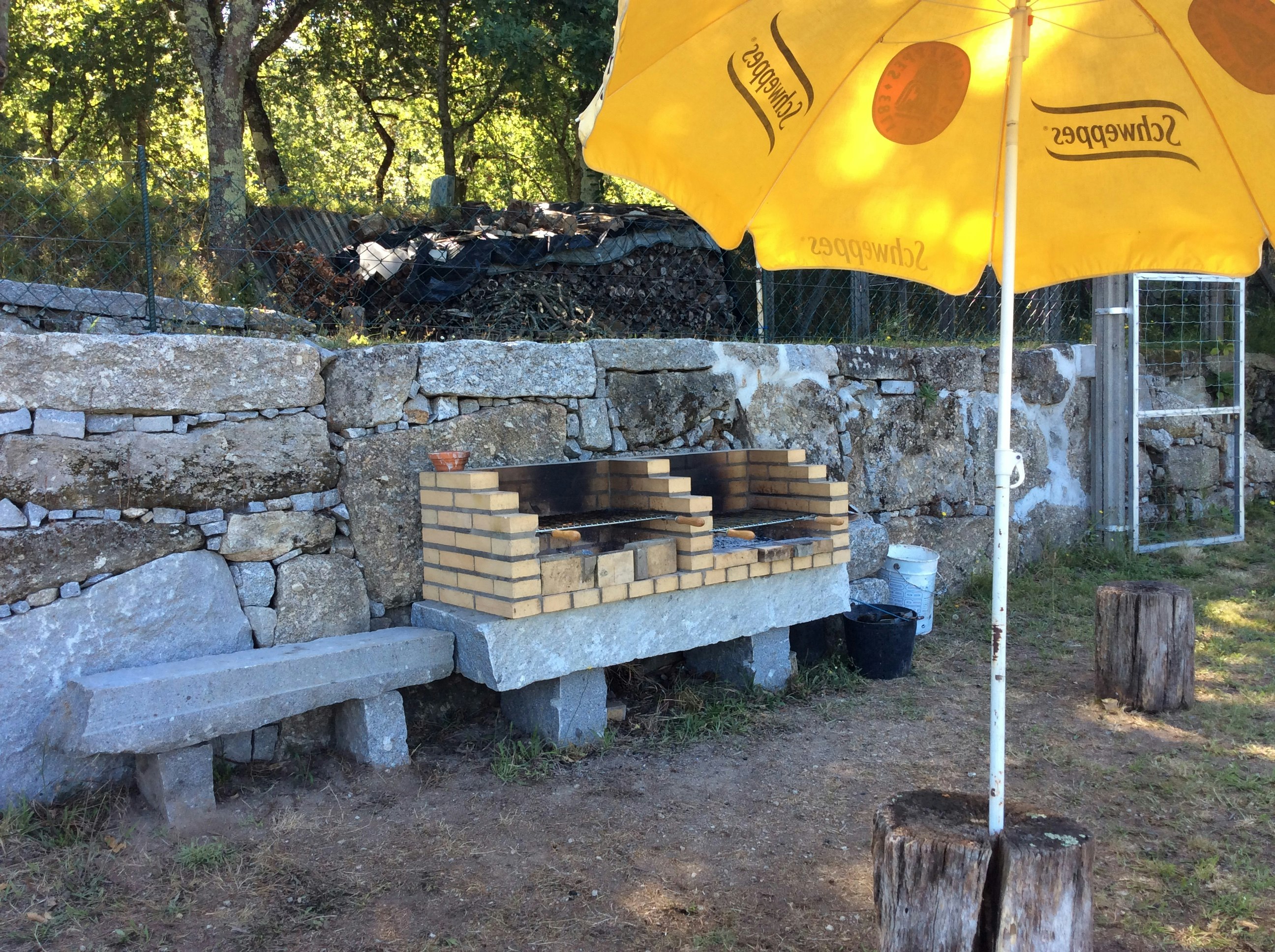 Grillplatz am Campingplatz mit Steinmauer, Holzscheiten und gelbem Sonnenschirm über einer Feuerstelle.
