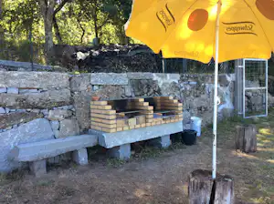 Grillplatz am Campingplatz mit Steinmauer, Holzscheiten und gelbem Sonnenschirm über einer Feuerstelle.