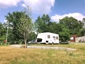 Wohnmobil auf einem Campingplatz auf einer Wiese, daneben ein Auto, Bäume und ein kleiner Schuppen im Hintergrund.
