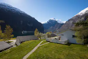 Campinganlage am See mit Blick auf schneebedeckte Berge, Rasen, Wege und Holzterrasse an einem weißen Gebäude.