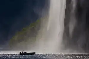 Boot auf dem See vor einem Wasserfall, Nebel und Felswand im Hintergrund, Sonnenlicht durch die Gischt.