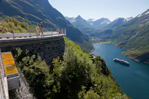 Aussichtsterrasse am Fluss im Tal mit Geländer, Wanderern und Blick auf Berge, Wald und ein Schiff auf dem Wasser.