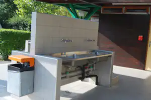 Außenküche am Campingplatz mit Spüle, Wasserhähnen, Fliesenboden und Abfallbehälter neben einer überdachten Terrasse.
