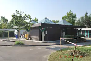 Camping-Unterstand mit Metall-Dach, grünen Stützen und Sitzbank, im Hintergrund Bäume und Mülltonnen am Weg.