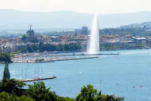 Blick auf einen See mit Fontäne, Booten und Uferpromenade, dahinter eine Stadt mit Türmen und Gebäuden.