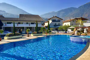 Schwimmbecken mit Liegen am Campingplatz, dahinter mehrstöckige Gebäude und Berge in der Ferne.