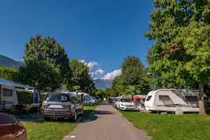 Campingplatz-Gasse mit geparkten Autos und Wohnwagen, Rasenstreifen und Bäumen entlang des Weges, im Hintergrund Berge.