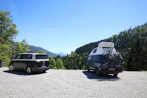 Zwei Wohnmobile auf einem Campingplatz-Parkplatz, im Hintergrund Berge und Nadelbäume, daneben ein Auto.