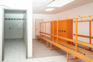 Duschraum mit gefliesten Wänden, zwei Wasserhähnen und orangefarbenen Bänken vor Spinden im Umkleidebereich.