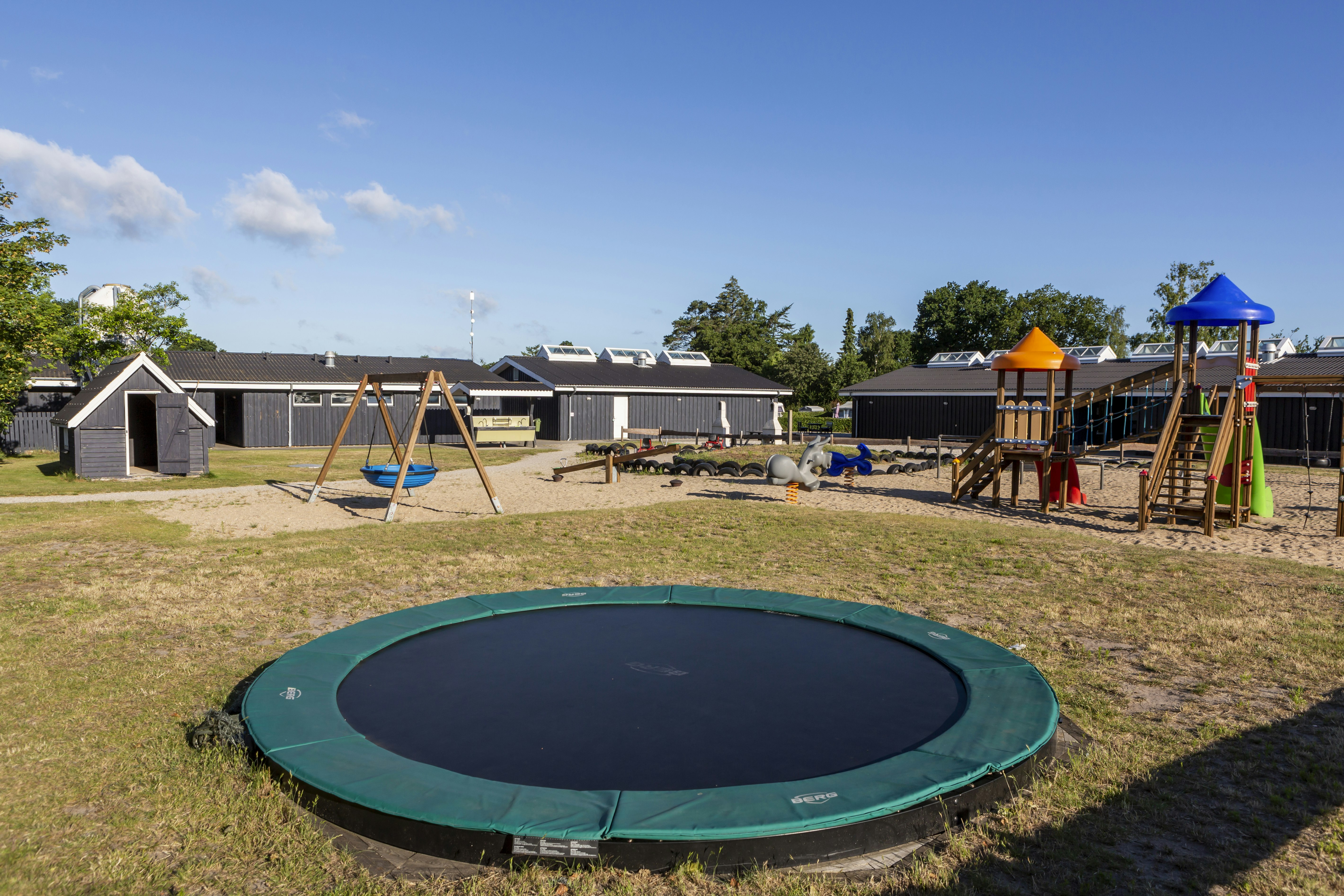 Trampolin auf dem Rasen vor Ferienhäusern und Wohnwagen, mit Spielgerüst und Sonnenschirm im Campingbereich.