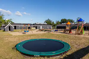 Trampolin auf dem Rasen vor Ferienhäusern und Wohnwagen, mit Spielgerüst und Sonnenschirm im Campingbereich.