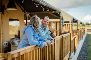 Paar auf der Holzterrasse eines Wohnwagens im Campingbereich, hält Tassen und sitzt am Geländer.